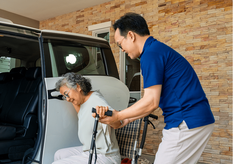 An older woman with a walker gets help from a man as she exits a car in a driveway with a brick wall in the background.